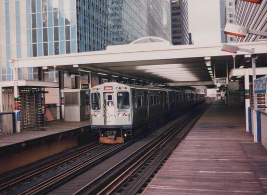 A silver and blue Chicago 'L' train, labeled 702, is stopped at an elevated station with a wooden platform and modern buildings in the background.