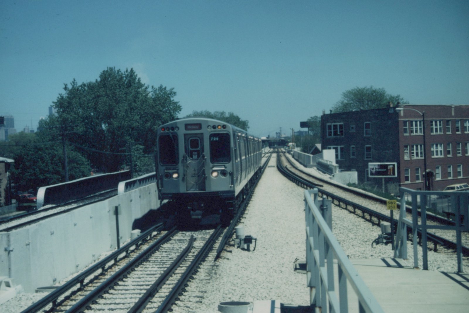 A silver and blue Chicago 'L' train, car 766, is shown approaching on elevated tracks with a clear blue sky and city buildings in the background.
