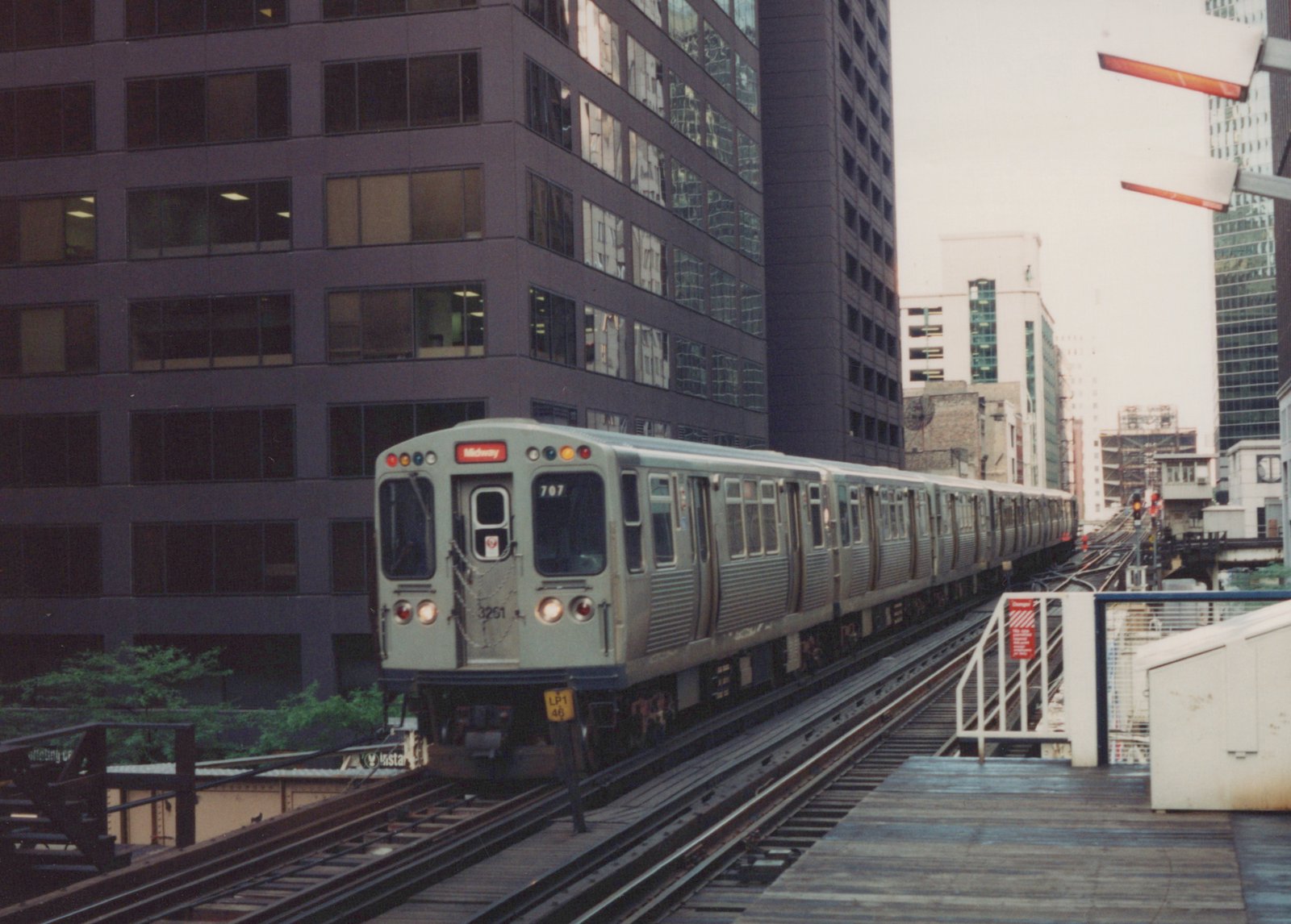 A silver Chicago 'L' train with the number 707 and 'Midway' destination sign travels on an elevated track beside tall office buildings.