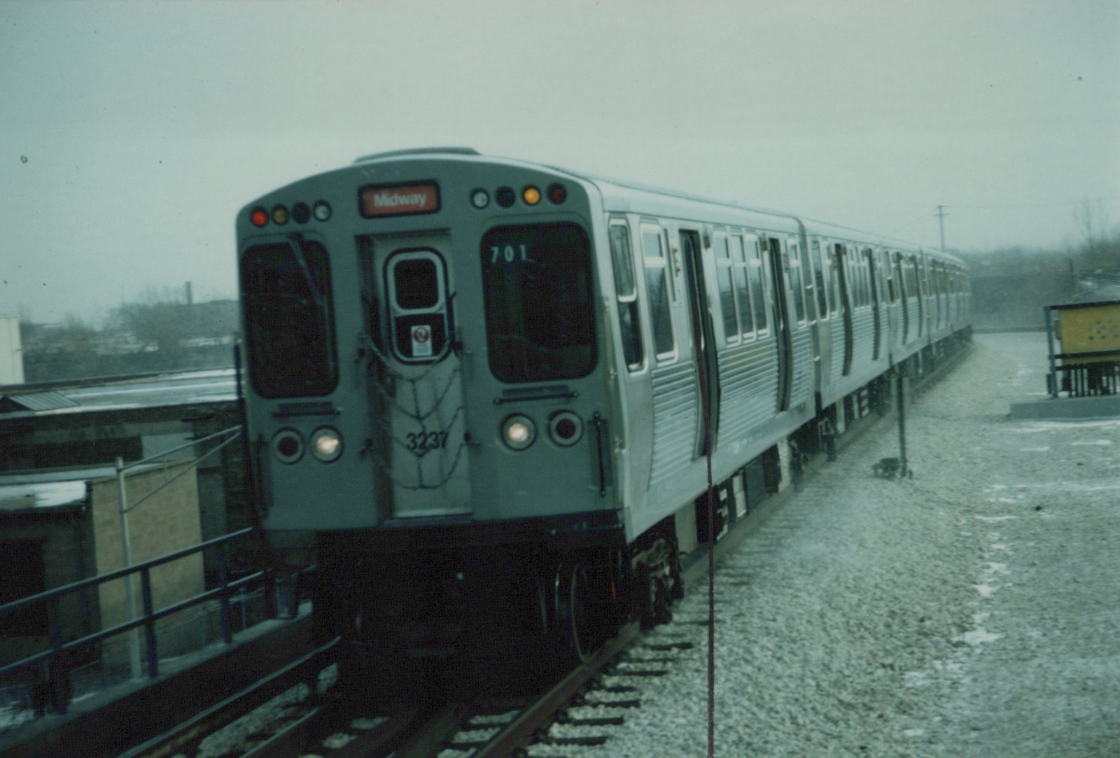 A silver Chicago Transit Authority rapid transit train is parked on an elevated track in winter, with snow on the ground and its destination sign lit for Midway.