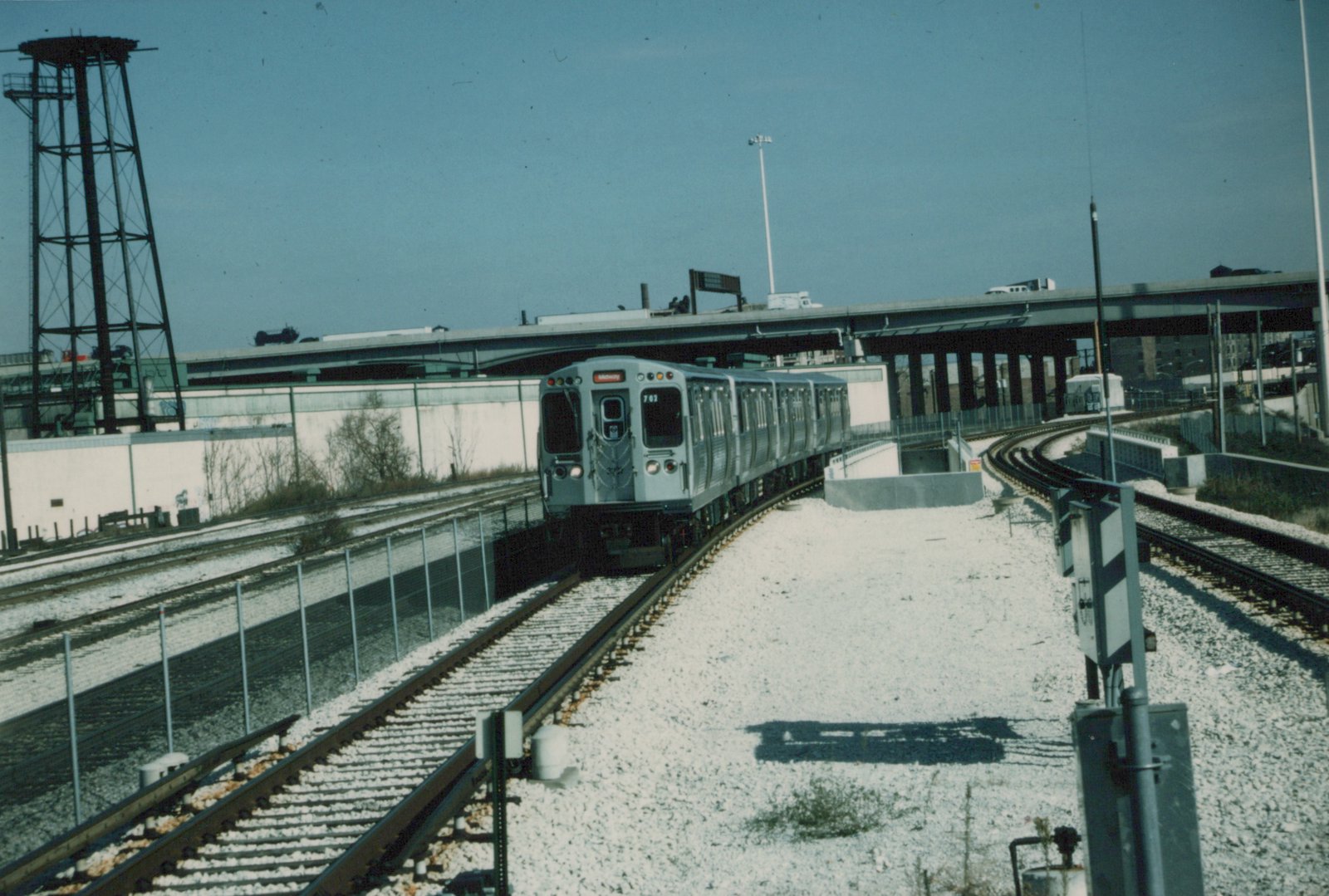 A silver Chicago Transit Authority rapid transit train, numbered 702, travels along a curved track in winter, with snow on the ground and an elevated highway and a water tower in the background.