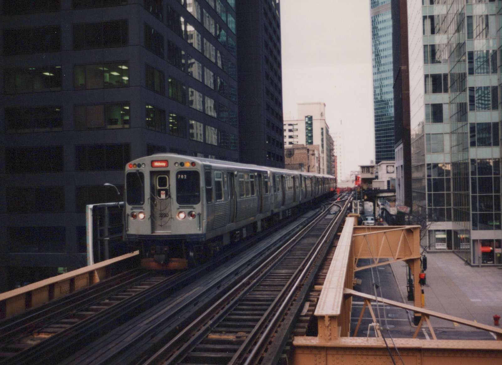 A silver CTA train numbered 3220 is shown on elevated tracks between tall buildings on an overcast day.