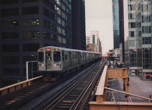 A silver CTA train numbered 3220 is shown on elevated tracks between tall buildings on an overcast day.