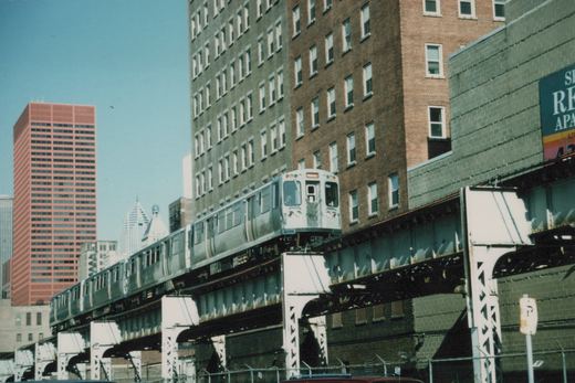 A silver Chicago 'L' train, number 704, travels on an elevated track with tall buildings and a blue sky in the background.