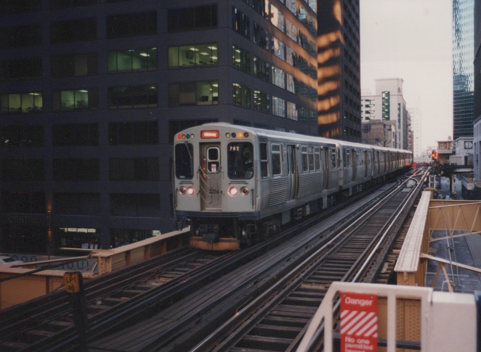 A silver Chicago rapid transit train on elevated tracks at dusk, with city buildings in the background and the number 3226 visible on the front.