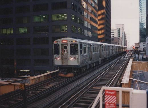 A silver Chicago rapid transit train on elevated tracks at dusk, with city buildings in the background and the number 3226 visible on the front.