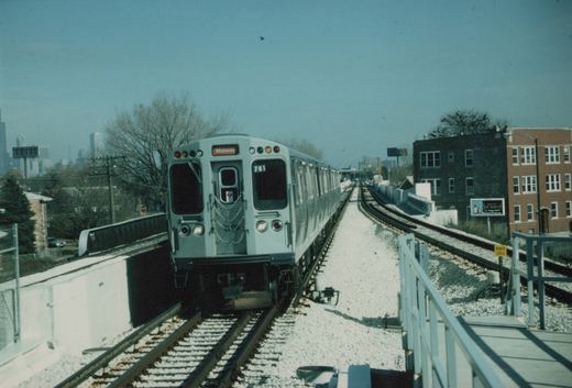 A silver Chicago Transit Authority train, number 701, on an elevated track with snow on the ground, traveling toward the Midway destination, with buildings and a city skyline in the background.