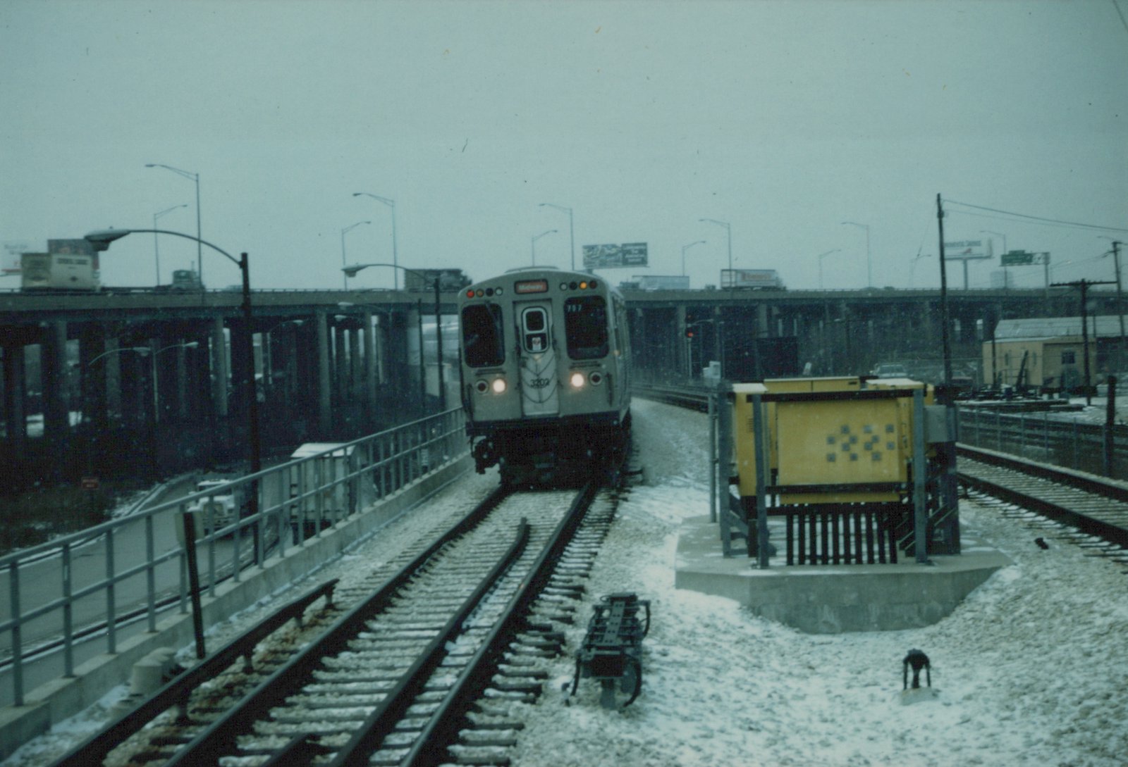 A silver CTA train with the number 3202 and a Midway destination sign is on elevated tracks with snow on the ground and an overpass in the background.