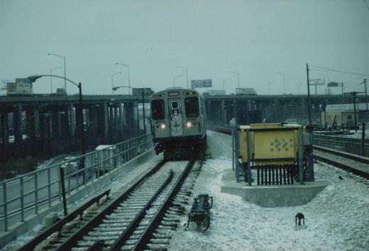 A silver CTA train with the number 3202 and a Midway destination sign is on elevated tracks with snow on the ground and an overpass in the background.