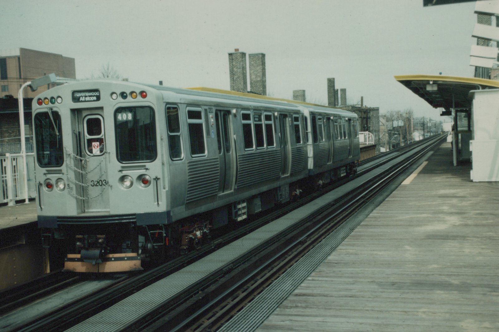 A silver CTA 2000-series rapid transit train with the number 3203 is stopped at an elevated station platform, with the destination sign reading 'Ravenswood' and the route number 408 displayed.