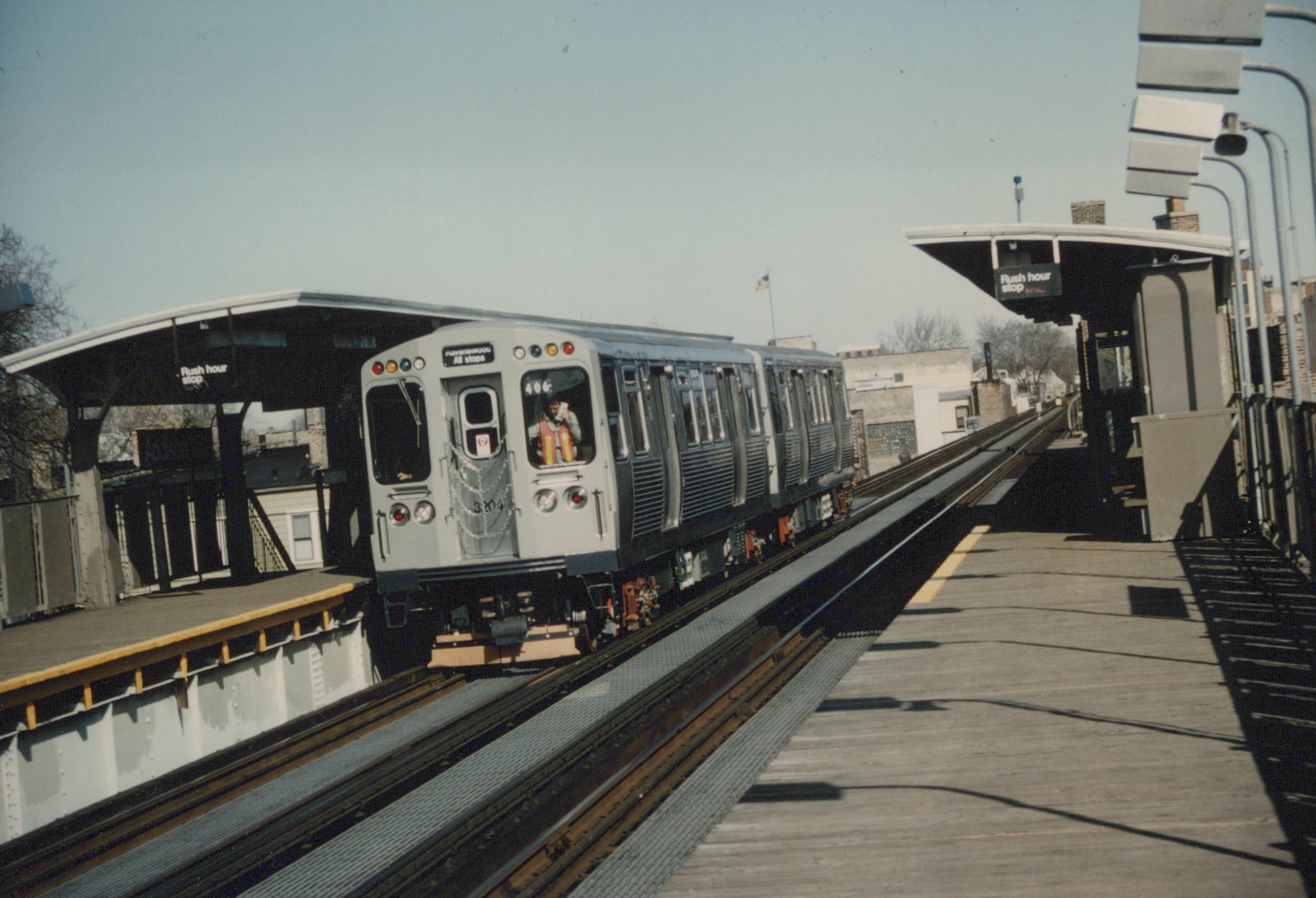 A silver rapid transit train with the number 3204 is stopped at an elevated station platform with a sign reading 'Rush hour stop'.