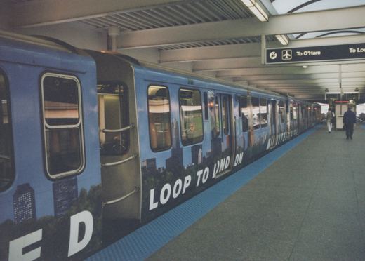 A blue rapid transit train with the number 3152 and an advertisement on its side is stopped at an indoor station platform with a few people walking nearby.