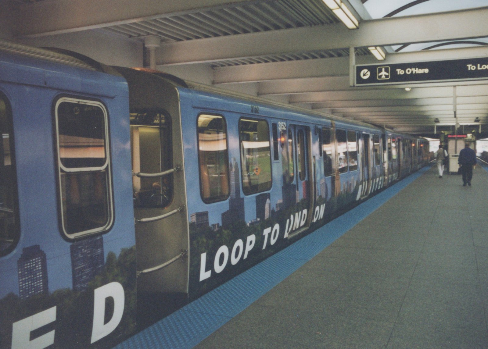 A blue rapid transit train with the number 3152 and an advertisement on its side is stopped at an indoor station platform with a few people walking nearby.