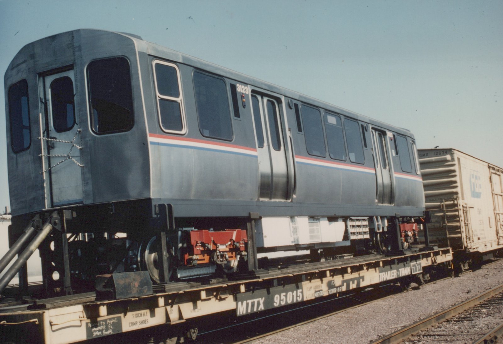 A stainless steel Chicago Transit Authority rapid transit car with fleet number 3123 is loaded on a flatcar, with other freight cars visible in the background in a sunny rail yard.