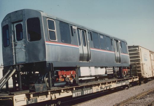 A stainless steel Chicago Transit Authority rapid transit car with fleet number 3123 is loaded on a flatcar, with other freight cars visible in the background in a sunny rail yard.
