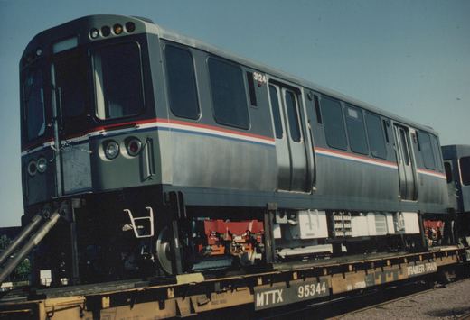 A silver Chicago Transit Authority rapid transit car with red and blue stripes, numbered 3124, sits on a flat railcar for transport on a sunny day.