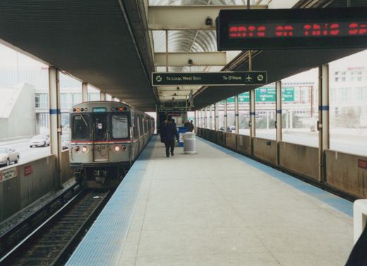 A silver CTA train with red and blue stripes, numbered 3113, is stopped at an elevated station platform with a few people walking on it. Signs indicate directions to O'Hare and the Loop.