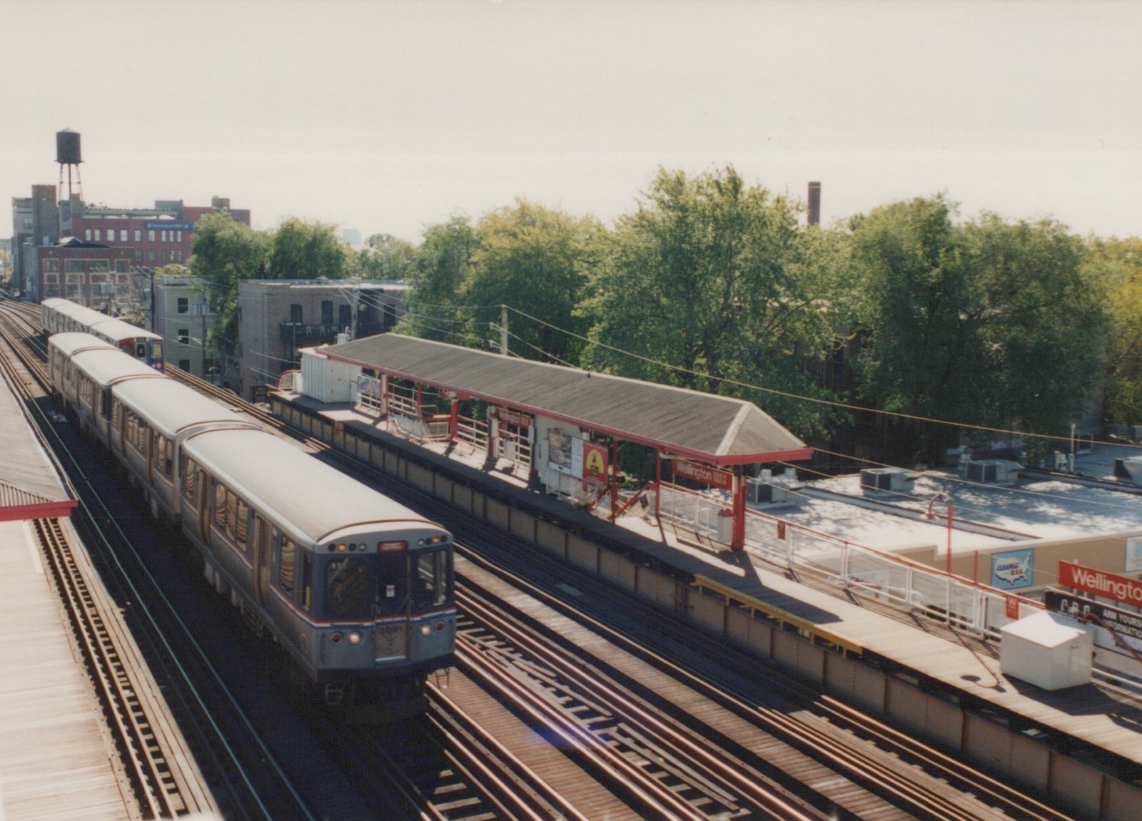 A blue and red CTA train with the number 3091 is moving on an elevated track beside a station platform labeled 'Wellington', with trees and buildings in the background.