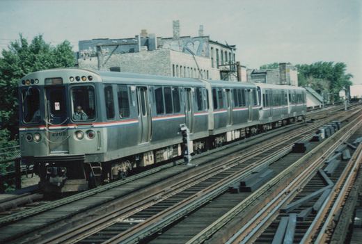A silver CTA train with red and blue stripes, numbered 2969, moves on elevated tracks with a building and trees in the background.