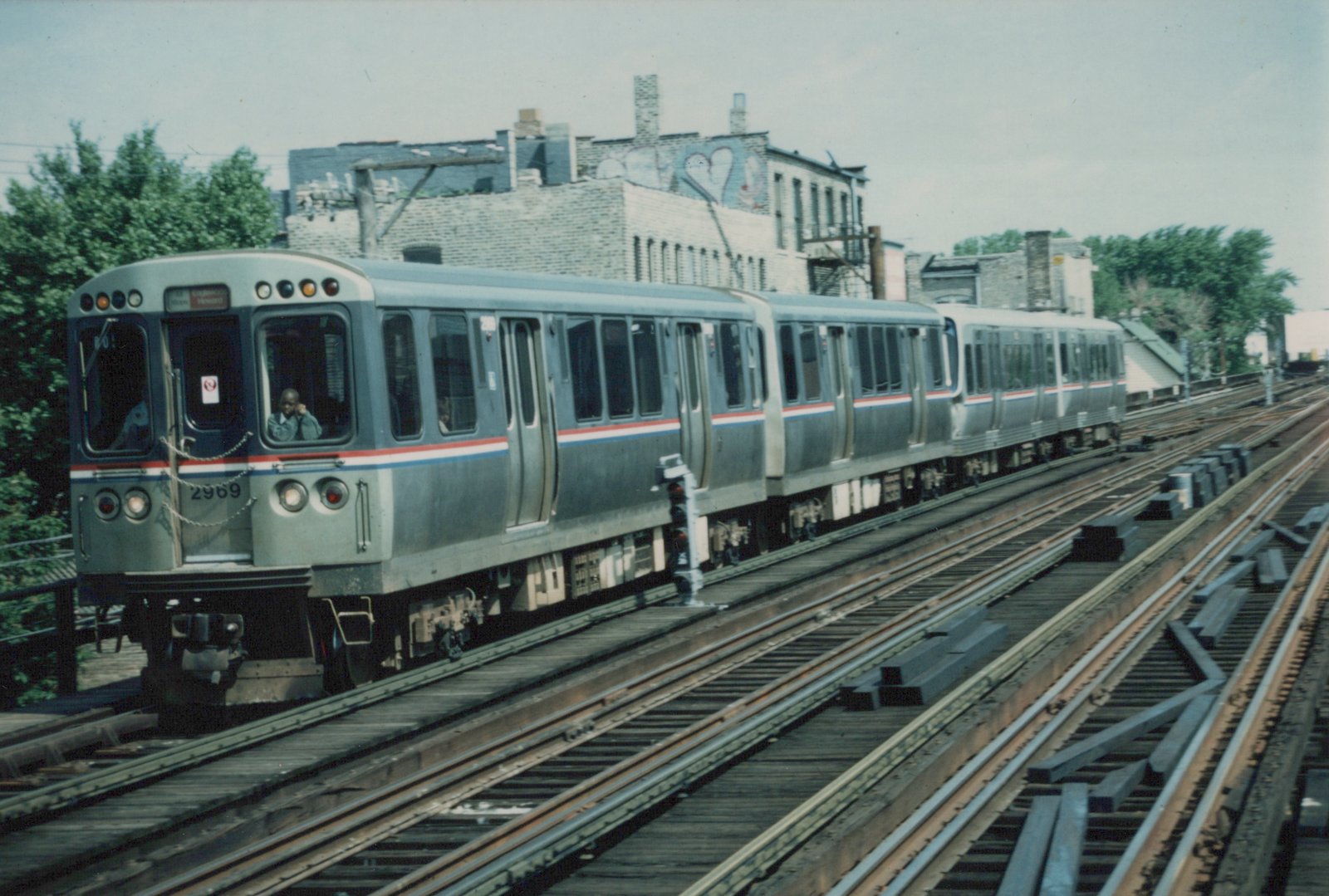 A silver CTA train with red and blue stripes, numbered 2969, moves on elevated tracks with a building and trees in the background.