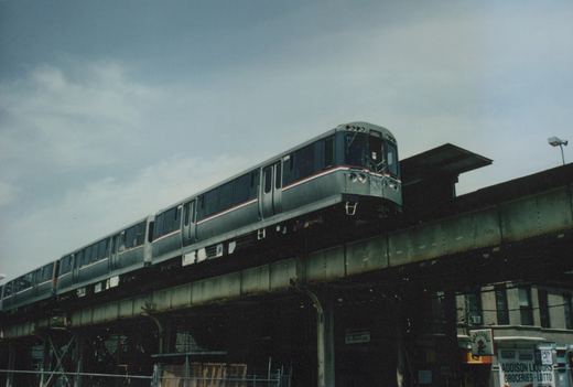 A silver CTA rapid transit train with red and blue stripes travels on an elevated structure above a street with storefronts, including one for Addison Liquors.
