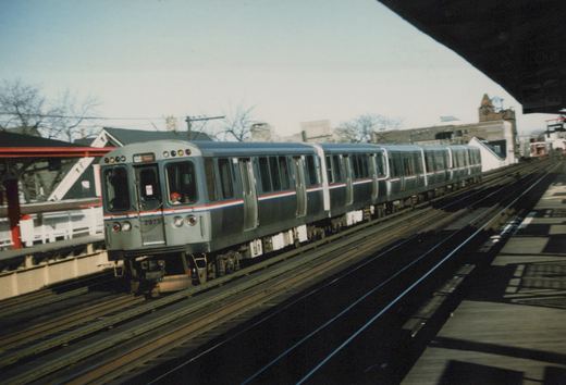 A silver and blue CTA train with the number 2926 on its front is moving along elevated tracks next to a station platform under a clear sky in winter.
