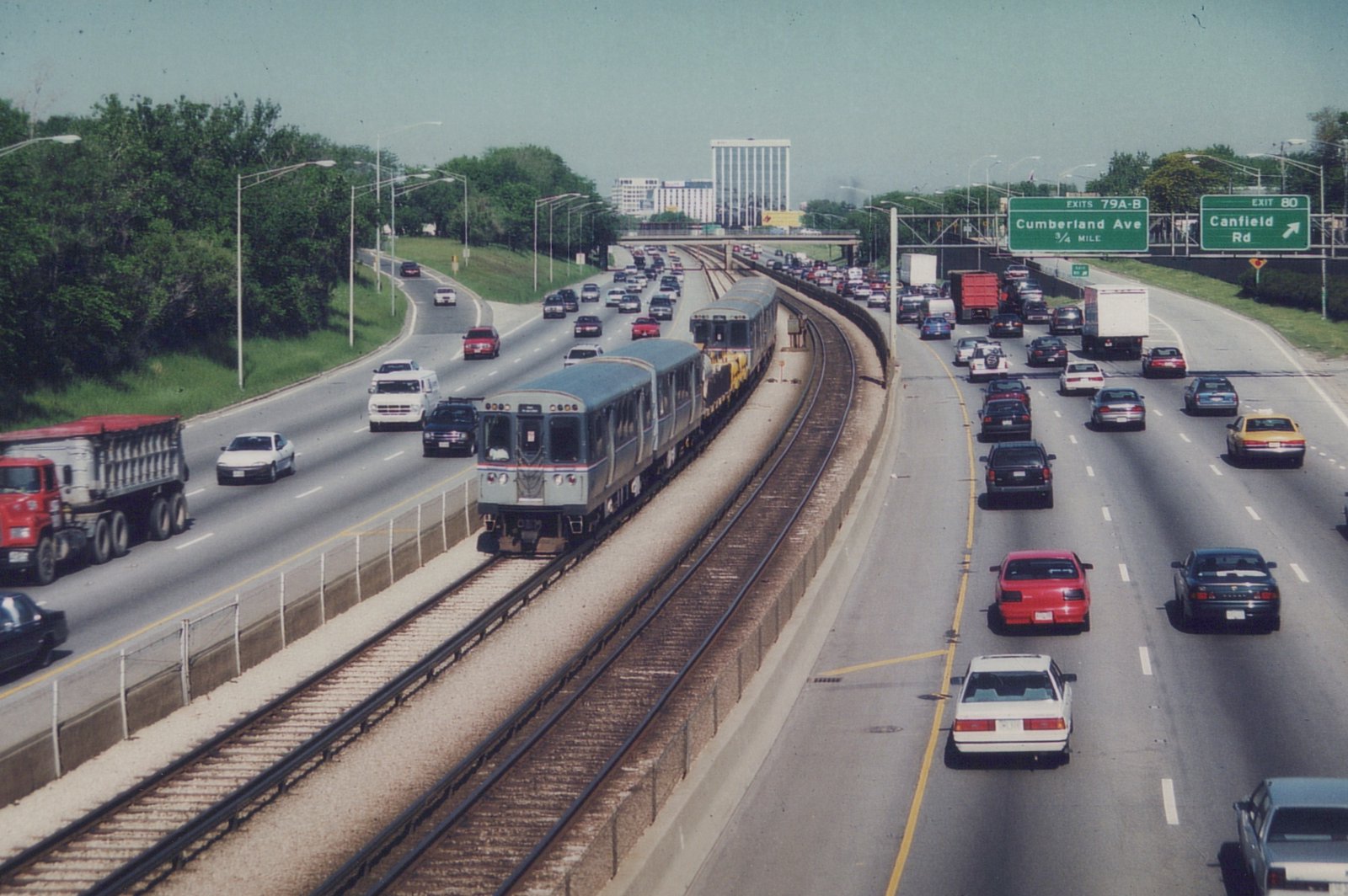 An elevated view of a gray CTA train with blue and red stripes moving through the median of a multi-lane highway, surrounded by cars and trucks on a sunny day.