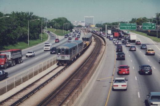 An elevated view of a gray CTA train with blue and red stripes moving through the median of a multi-lane highway, surrounded by cars and trucks on a sunny day.