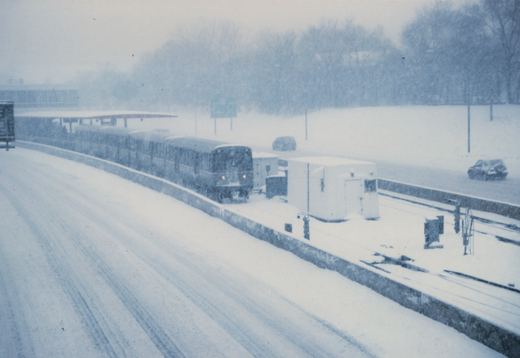 A train travels along tracks in the median of a snow-covered highway during a blizzard, with cars visible on the road and snow falling heavily.