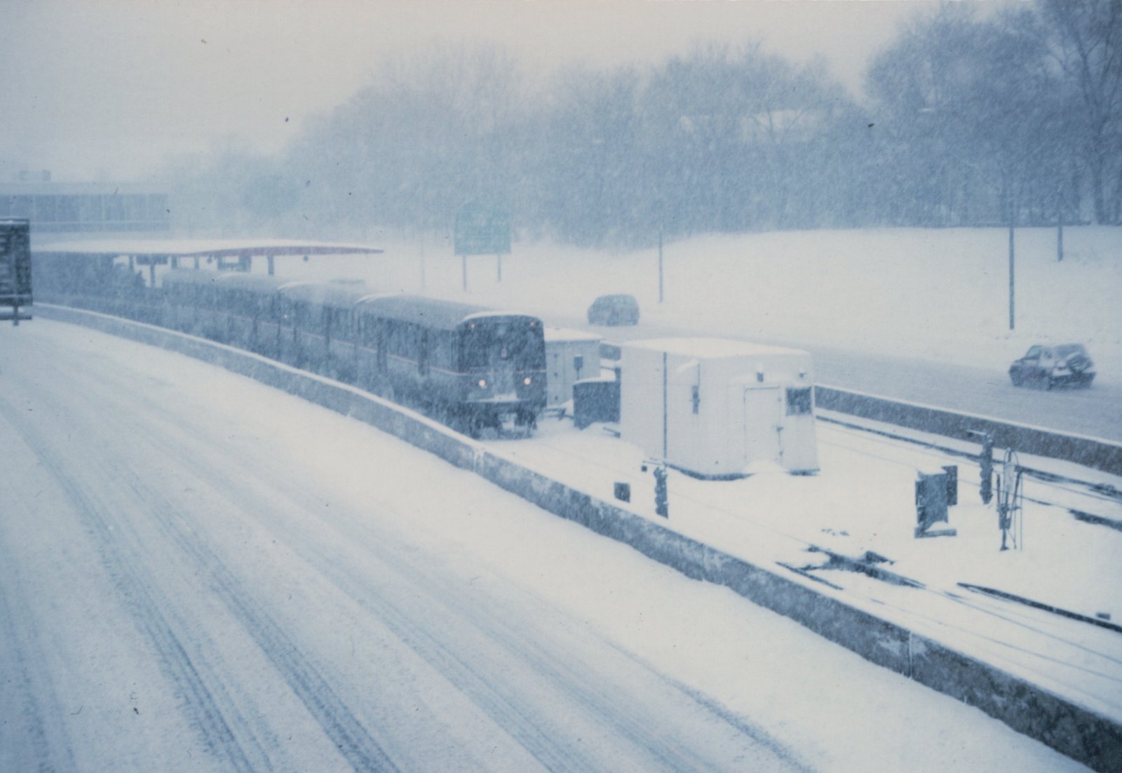 A train travels along tracks in the median of a snow-covered highway during a blizzard, with cars visible on the road and snow falling heavily.