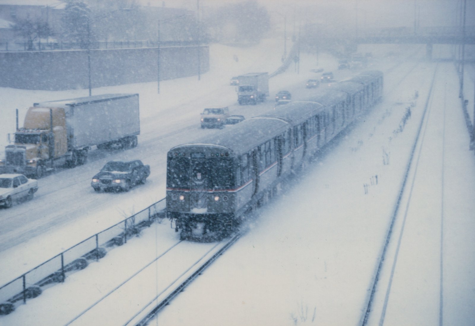 A Chicago 'L' train of the 2600 series moves through a snowstorm on a private right-of-way next to a highway with cars and trucks.