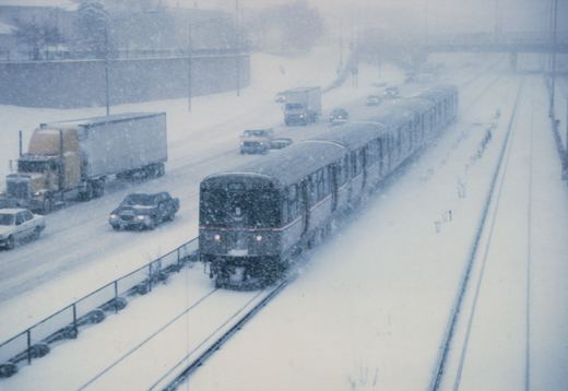 A Chicago 'L' train of the 2600 series moves through a snowstorm on a private right-of-way next to a highway with cars and trucks.