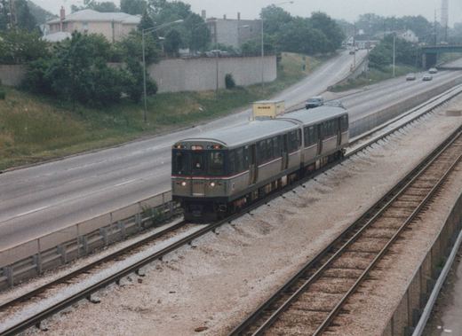 A silver Chicago 'L' train with the number 2209 travels on tracks in the middle of a multi-lane highway, with cars and a yellow Ryder truck visible on the road.