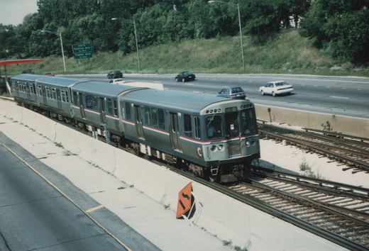A silver Chicago Transit Authority train with red and blue stripes, number 2204, moves on elevated tracks beside a multi-lane highway with cars, under a clear sky.