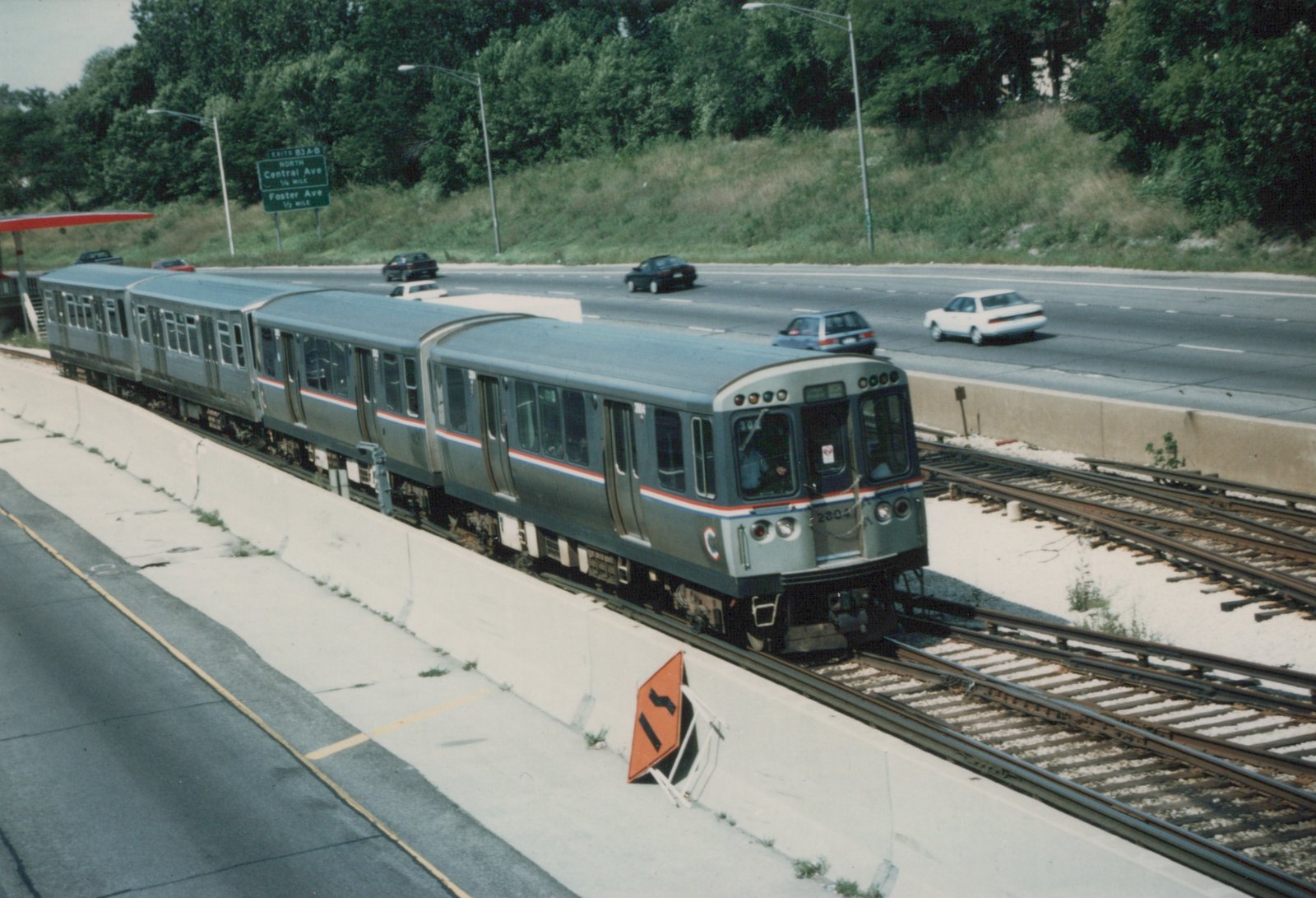 A silver Chicago Transit Authority train with red and blue stripes, number 2204, moves on elevated tracks beside a multi-lane highway with cars, under a clear sky.