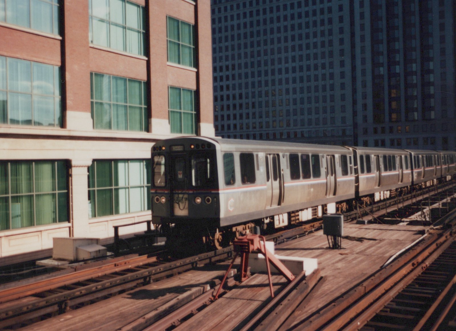 A silver CTA train with red and blue stripes, number 2771, travels on a wooden elevated track between tall city buildings on a sunny day.