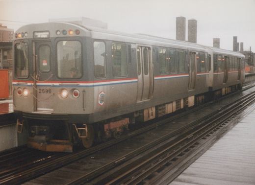 A silver rapid transit car with red, white, and blue stripes, numbered 2696, is stopped at an elevated station platform under an overcast sky.