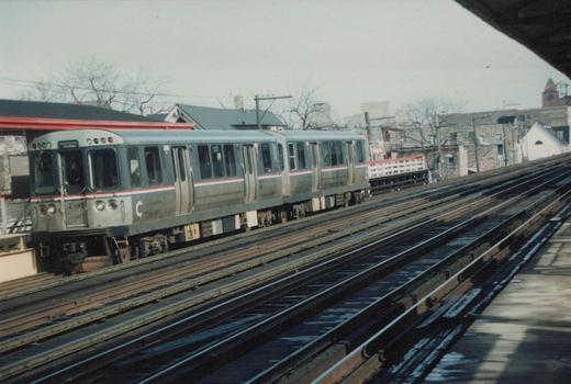 A silver Chicago Transit Authority rapid transit train with red and blue stripes is stopped at an elevated station, with the front showing the number 2702 and destination sign reading 'HARWOOD All stops'.