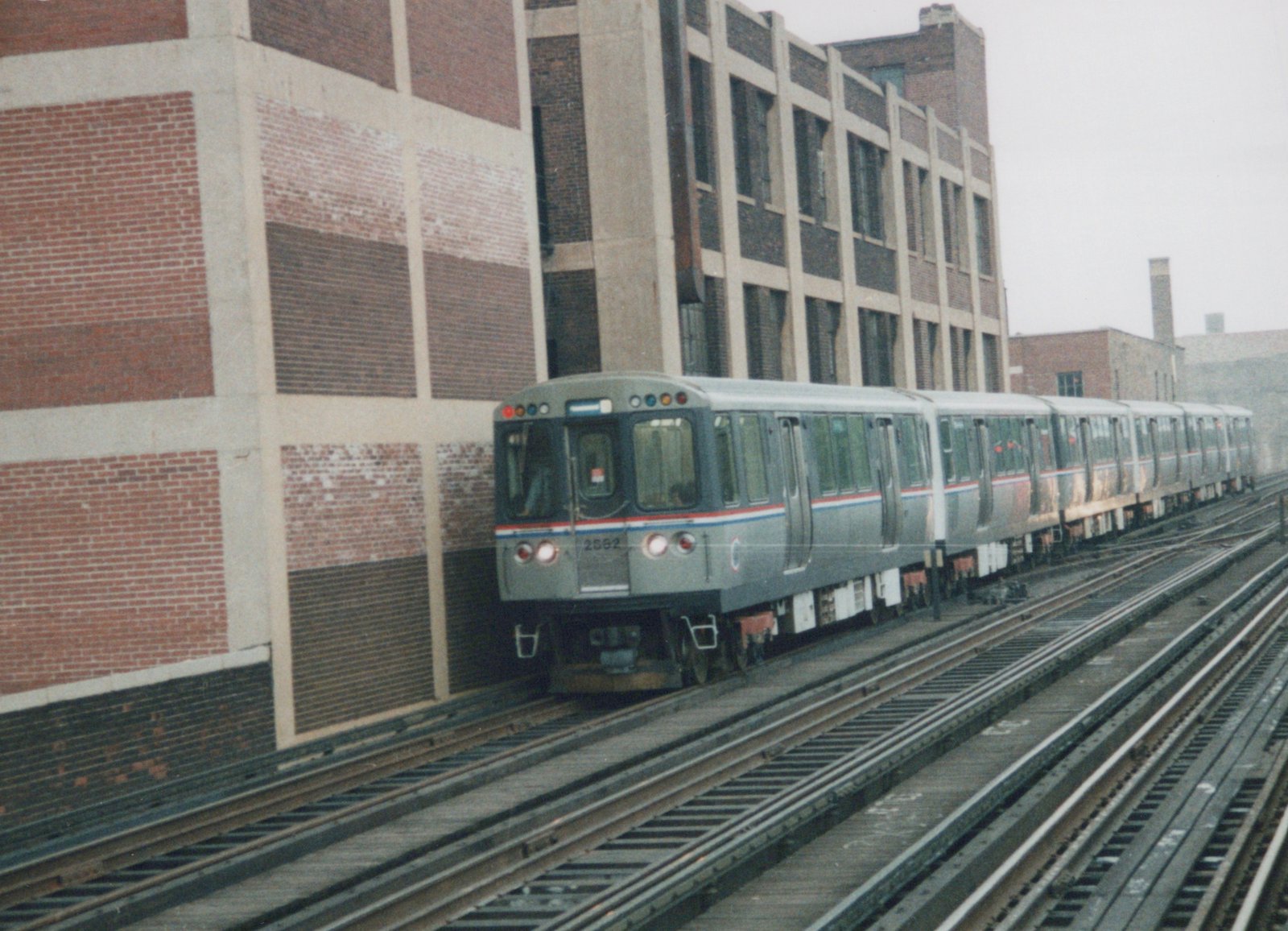 A gray CTA train with red and blue stripes, numbered 2562, on elevated tracks beside a large brick building on an overcast day.