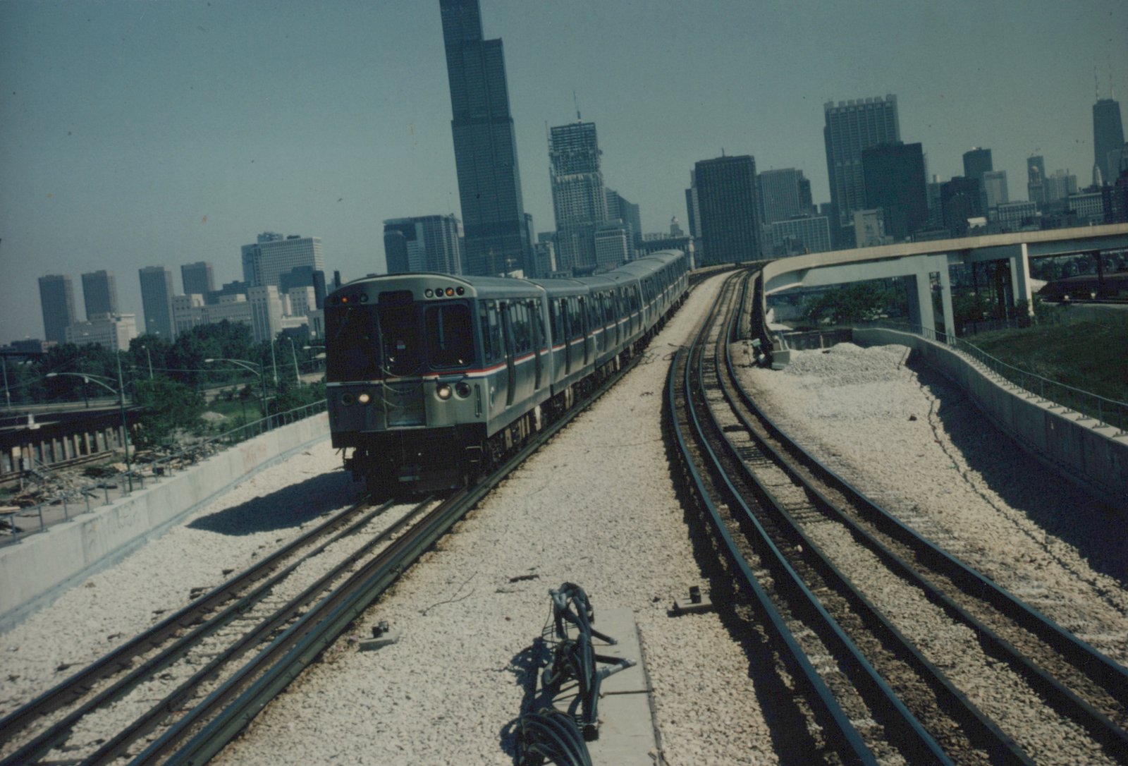 A silver train with red and blue stripes travels on an elevated track with the Chicago skyline behind it on a clear day.
