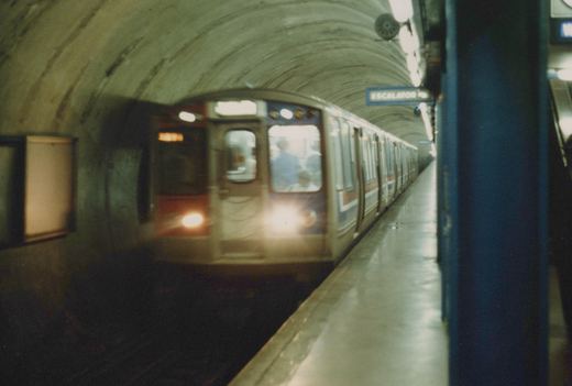 A blurred CTA 2000-series subway train moving through a concrete-arched station tunnel with a blue 'ESCALATOR' sign visible on the platform wall.