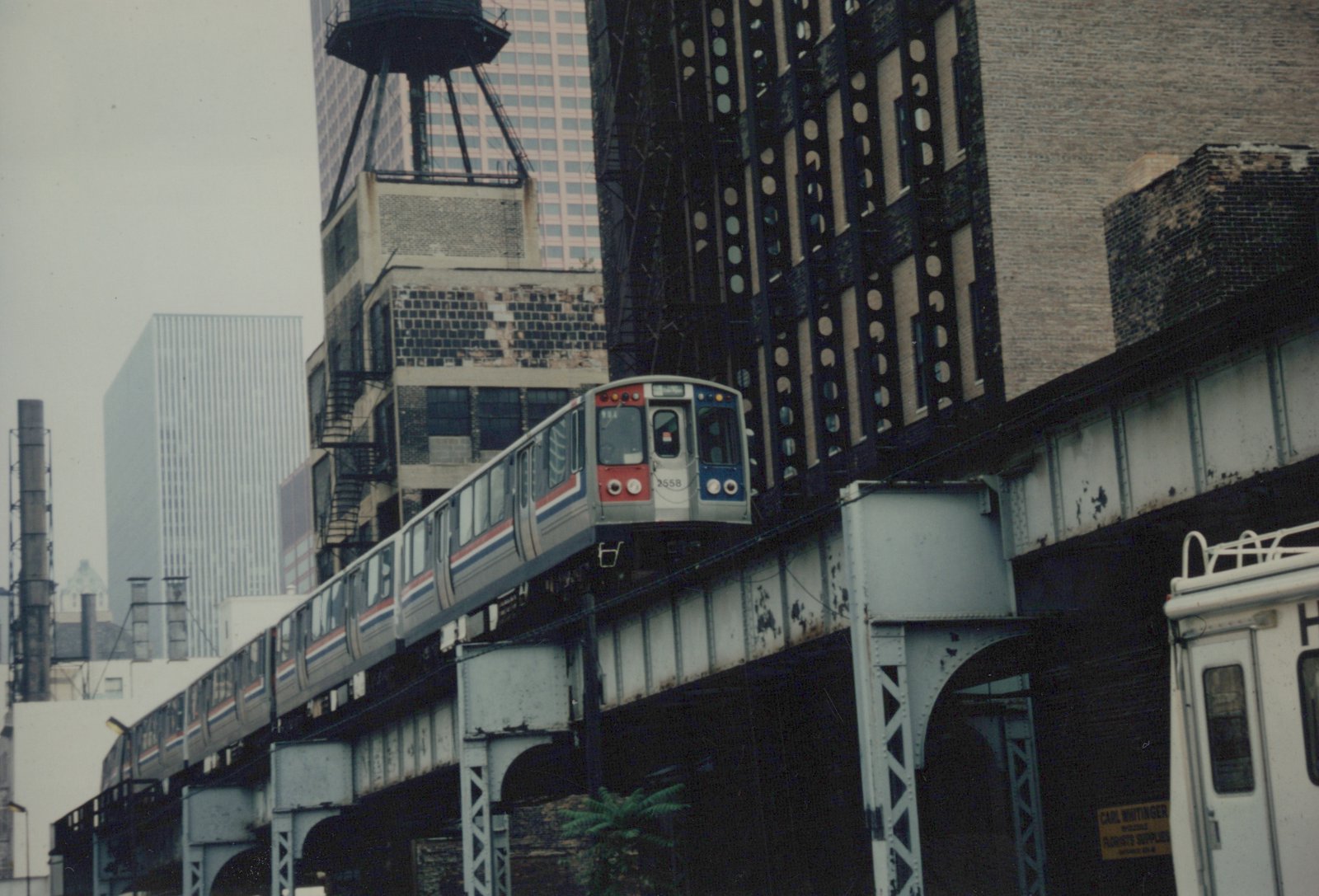 A silver Chicago 'L' train with red and blue stripes, numbered 2558, travels on an elevated track against a backdrop of city buildings on an overcast day.