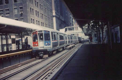 A silver, red, and blue rapid transit train with the number 2552 is stopped at an elevated station platform. The station has wooden decking and steel trusses, with tall buildings in the background.
