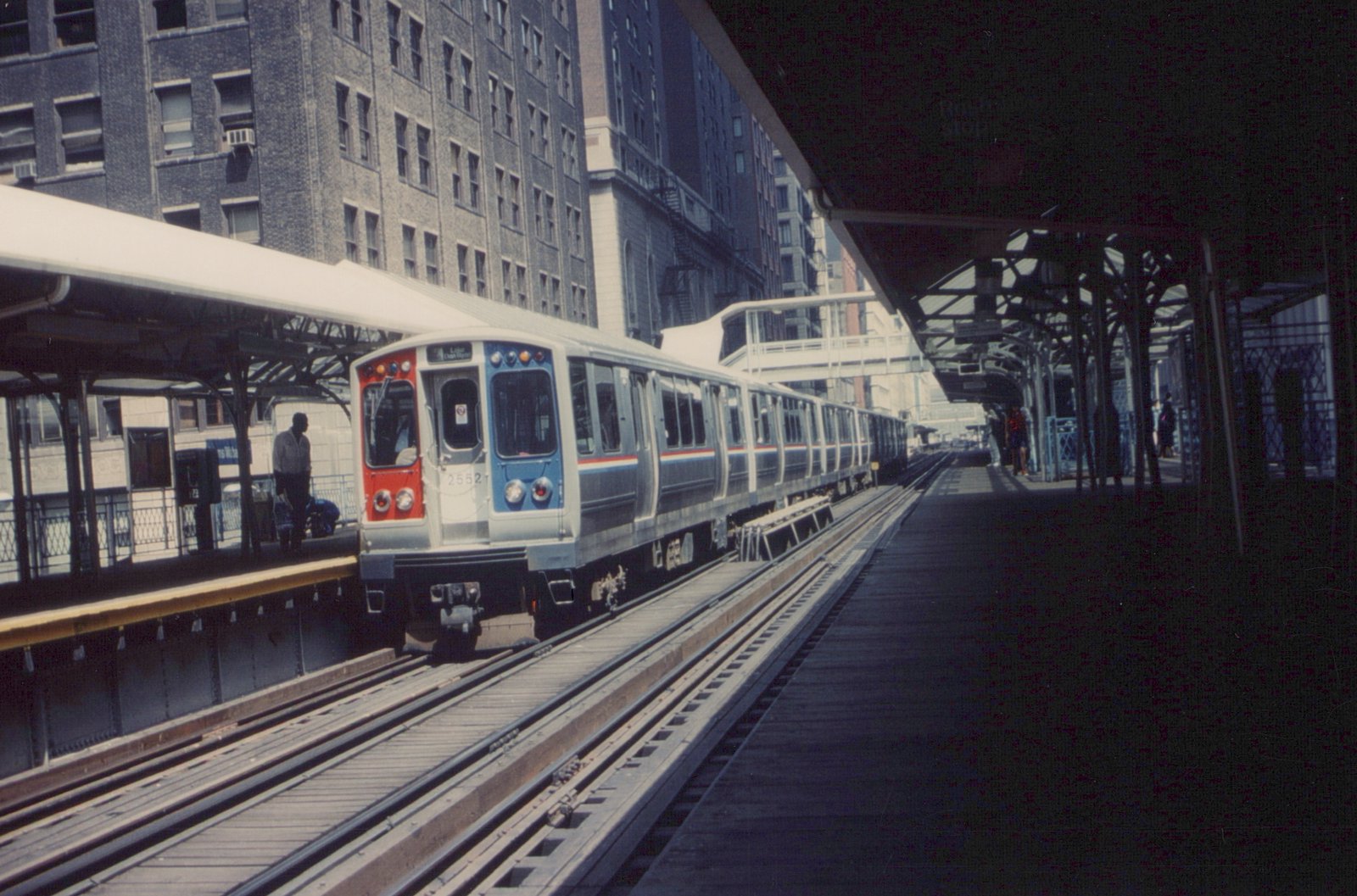 A silver, red, and blue rapid transit train with the number 2552 is stopped at an elevated station platform. The station has wooden decking and steel trusses, with tall buildings in the background.