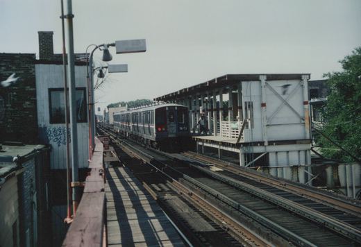 A silver and red train with the number 2691 on the front is moving on elevated tracks toward a platform with a person standing on it.