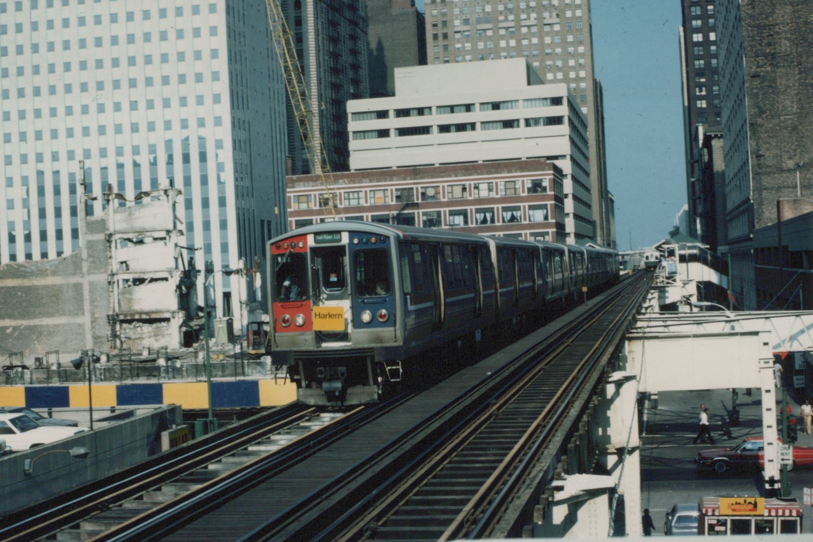 A blue and silver rapid transit train with 'Harlem' on the front sign moves along an elevated track in a city with tall buildings and a clear sky.