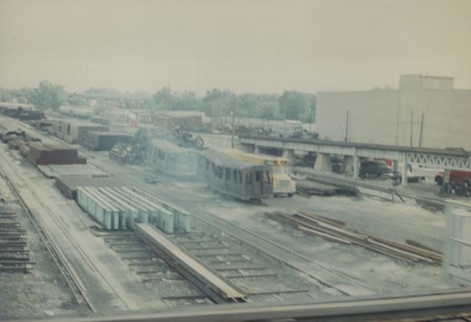 A gray rapid transit train on tracks in a large rail yard with freight cars, a yellow work truck, and a steel bridge in the background.