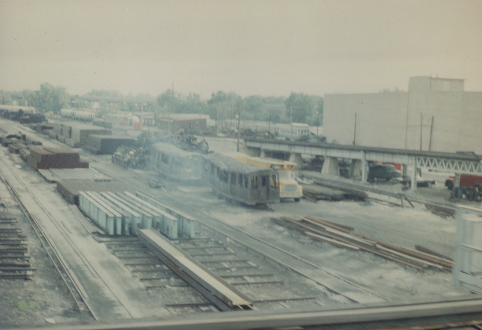 A gray rapid transit train on tracks in a large rail yard with freight cars, a yellow work truck, and a steel bridge in the background.