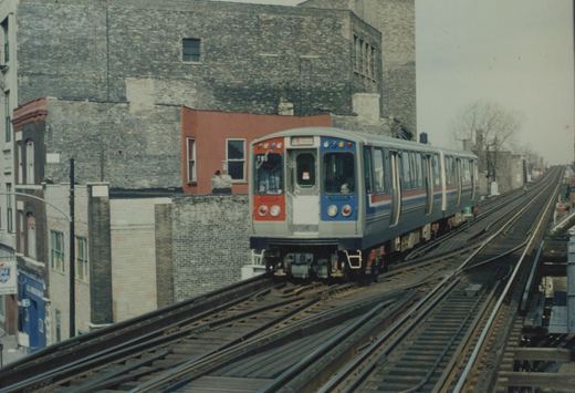 A blue, red and silver CTA train car marked 203 on elevated tracks next to brick buildings under an overcast sky.