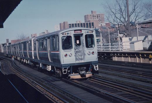 A silver and blue Chicago Transit Authority rapid transit train with the number 2501 on the front, on elevated tracks with buildings in the background and no leaves on the trees.